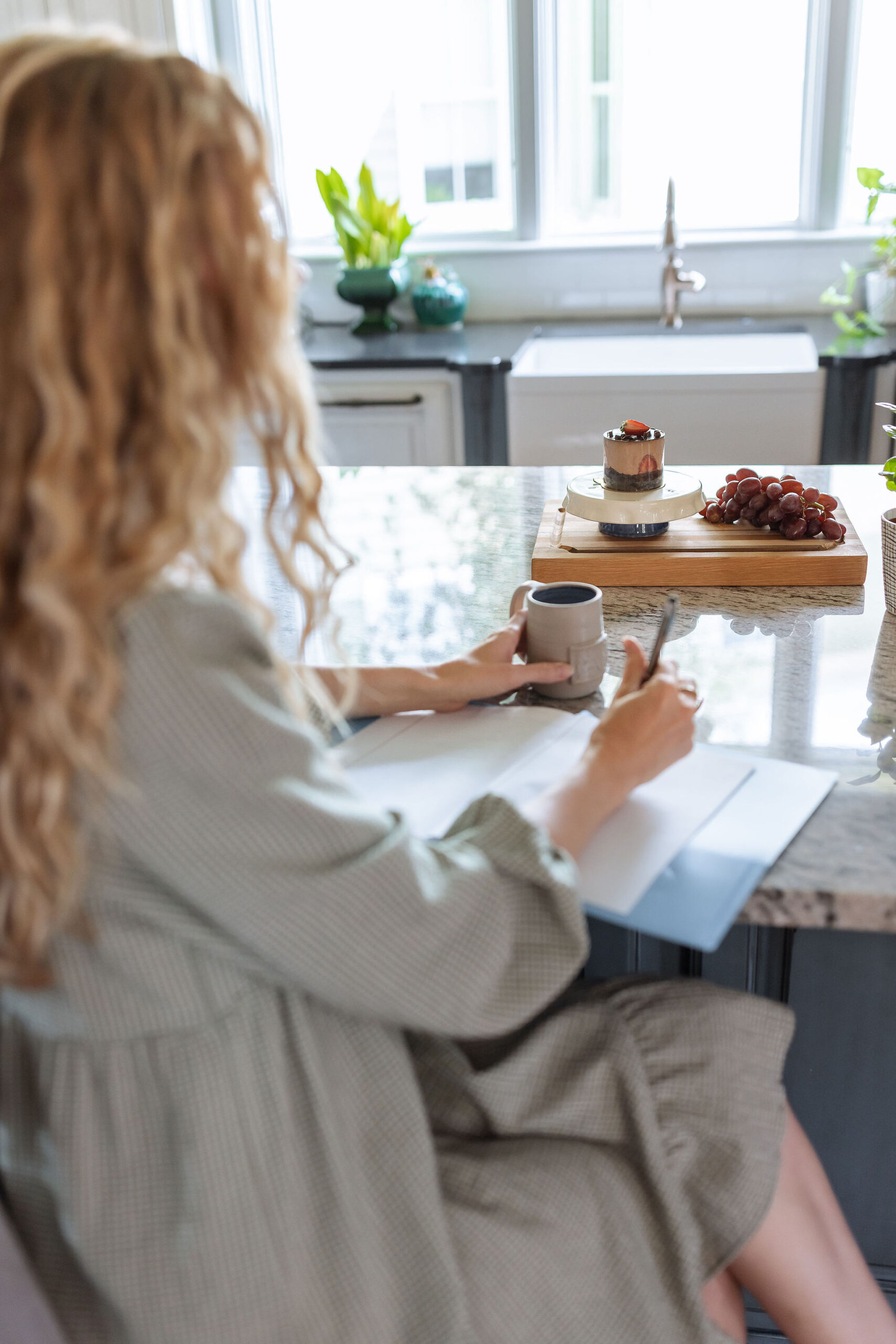 Board of grapes and chocolate cake on kitchen bar with woman writing in a blue notebook while holding a coffee cup as she helps clients feel calm with food and in their bodies